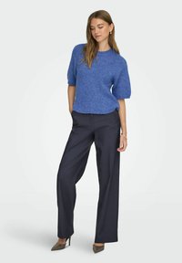 Modrý pletený svetr s krátkými rukávy, doplněný tmavě šedými širokými kalhotami a botami na vysokém podpatku. Hladká textura a volnější střih.