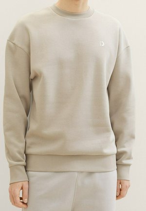 Sudadera beige con cuello redondo y pantalones a juego, usados por una persona que está de pie frente a un fondo claro y liso.