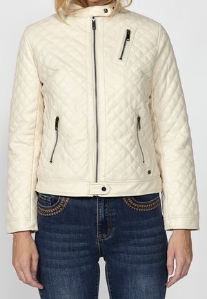 Kvinde iført en cremefarvet quiltet jakke med lynlåslommer og mørkeblå jeans med brune broderede detaljer på lommerne.