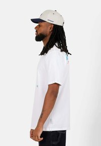 Witte T-shirt met korte mouwen en grafische ontwerpen in blauw en roze. Zwarte broek, gedragen met een gestreepte pet met een donker klepje.