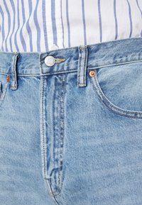 Närbild av ett ljusblått denimjeans midjeband och framfickan med kopparnitar, bärs tillsammans med en inknäppt vit skjorta med vertikala blå ränder.