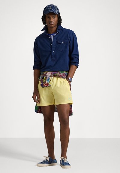 Modelo vestindo uma camisa de colarinho azul-marinho de mangas longas, calções amarelos e sapatos azuis com padrão. Uma camisa de padrão xadrez está amarrada à volta da cintura.