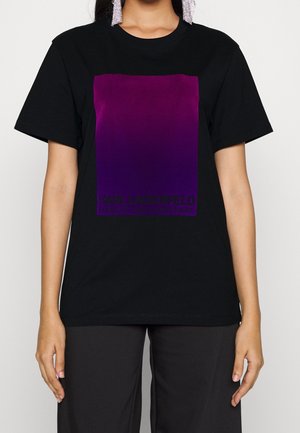 Persoon draagt een zwart T-shirt met korte mouwen met een groot paars vierkant en de tekst Karl Lagerfeld op de voorkant, gecombineerd met een zwarte broek.