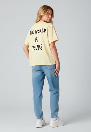 T-shirt di cotone gialla con la scritta "IL MONDO È TUO" stampata in grassetto nero sulla schiena. Abbinata a pantaloni di jeans azzurri chiari e sneakers bianche.