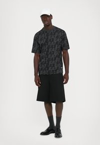 Man wearing black patterned EA7 T-shirt, black wide shorts, gray socks, black loafers, and white cap standing against white background.