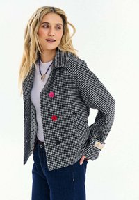 Žena s dlhými blond vlasmi, oblečená v čierno-bielom kabáte so vzorom houndstooth a farebnými gombíkmi, bielou košeľou, modrými džínsami a náhrdelníkom z korálok.