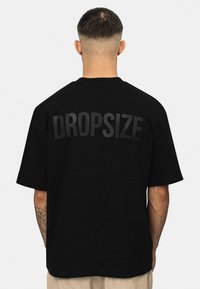 Sort t-shirt med korte ærmer, der har stor "DROPSIZE" tekst i mat finish på ryggen. Lavet af blødt bomuldsstof.