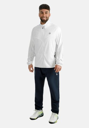 Mann steht lächelnd da, trägt eine weiße Jacke mit Reißverschluss, marineblaue Jogginghose und weiße Sportschuhe mit schwarzen und gelben Akzenten.