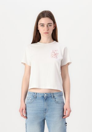 T-shirt blanc court en coton avec un col rond. Présente un design graphique rose sur la zone gauche de la poitrine. Porté avec un jean bleu clair.