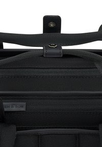 Gros plan sur un sac en tissu texturé noir montrant une fermeture éclair, deux sangles noires tissées et un bouton-pression.