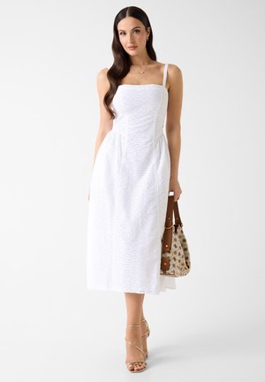 Ženska z dolgimi temnimi lasmi v beli midi obleki brez rokavov, z zlatimi sandali s trakovi in držo vzorčaste torbice na enobarvni podlagi.