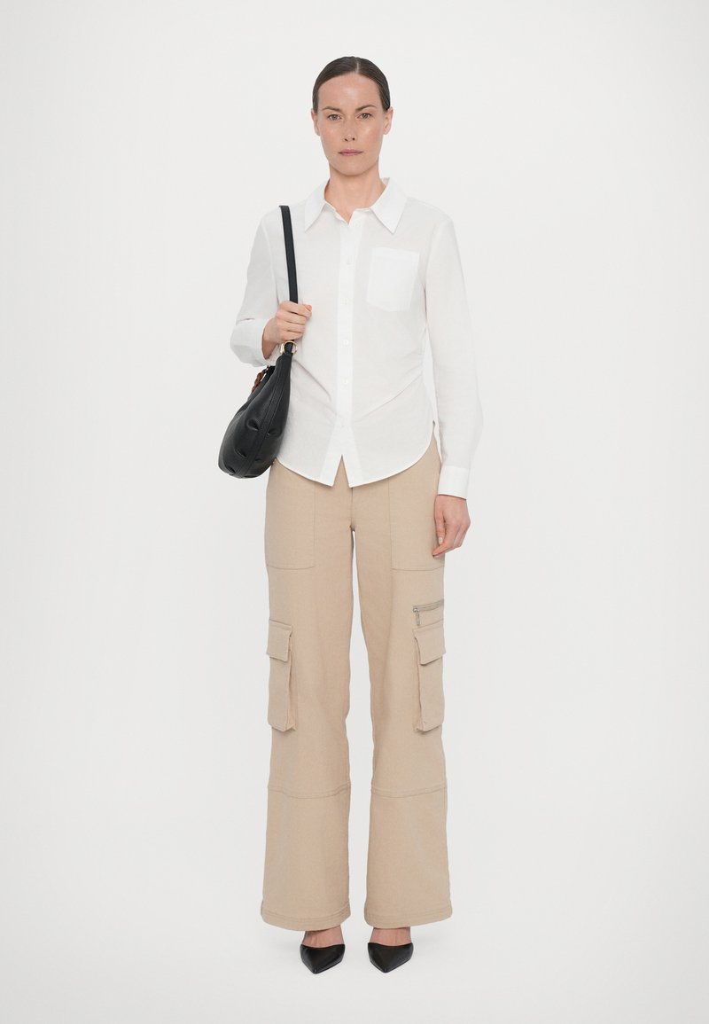 White long-sleeve button-up shirt with a chest pocket, paired with beige cargo pants featuring large pockets and a zippered pocket. Black handbag and heels.