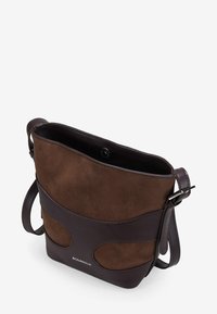 Bolso cruzado de ante y cuero marrón con textura suave, parte superior abierta y cierre metálico a presión. Presenta acentos de diseño curvado.