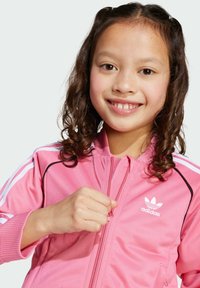 Vaaleanpunainen urheilujacketti, jossa on valkoinen Adidas-logo, vetskari edessä, ribattu hihansuu ja kolme valkoista raitaa hartioiden kohdalla. Pehmeä, sileä kangas.