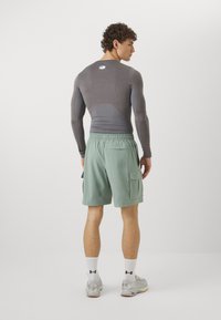 Sportinė apranga, kurią sudaro prigludusi pilka ilgomis rankovėmis marškiniai ir šviesiai žali šortai su šoninėmis kišenėmis ir elastingu juosmeniu.