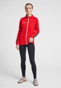 Rode sportjack met volledige rits, gestructureerde stof, zwarte patronen op de mouwen, gecombineerd met zwarte leggings en grijze schoenen.