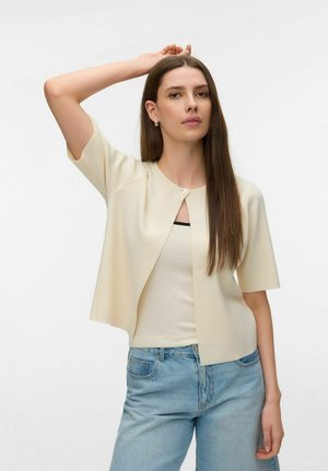 Hosszú barna hajú nő, krémszínű rövid ujjú kardigánt visel top felett és világoskék farmert, egyik karját felemelve pózol.