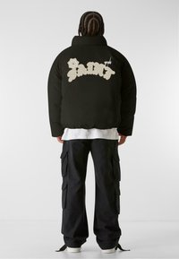Chaqueta de plumas negra con un gran gráfico en crema "SAINT" en la parte posterior, que presenta un diseño floral. Llevada sobre una camisa blanca y pantalones cargo negros.
