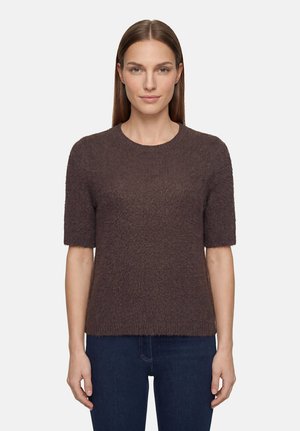 Femme aux cheveux bruns et raides porte un pull marron à manches courtes en tissu pelucheux et un jean bleu foncé, debout devant un fond blanc uni.