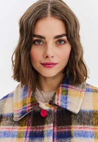 Vlněný kostkovaný kabát s kombinací žluté, modré a hnědé barvy, s růžovým knoflíkem a širokým límcem. Měkká textura a teplý design.