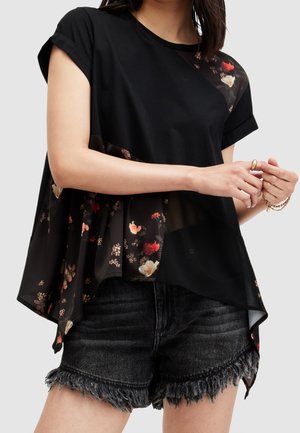 Woman wearing a black floral asymmetric blouse with rolled sleeves and dark frayed denim shorts, hands held near waist.