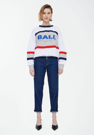 Sweatshirt blanc et gris avec des rayures bleues et rouges portant le logo "BALL", associé à un jean en denim bleu foncé, avec des ourlets retroussés et des chaussures noires à bout pointu.