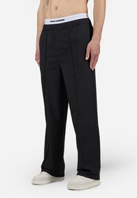 Pantalons noirs avec un design plissé, une taille élastique arborant le logo "PEGADOR" et un tissu lisse, texturé. Associés à des chaussures de couleur claire.