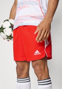 Piros sportnadrágok könnyű anyagból, fehér oldalsávokkal és Adidas logóval, fekete-fehér csíkos zoknikkal párosítva.