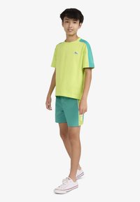 Φωτεινό πράσινο κοντομάνικο πουκάμισο με μπλε λεπτομέρειες και λογότυπο, συνδυασμένο με τιρκουάζ σορτς που έχουν ανοιχτοπράσινη πλευρική λωρίδα. Λευκά αθλητικά παπούτσια.