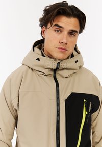 Beige wasserdichte Jacke mit einem schwarzen Akzentpaneel, verfügt über eine Reißverschlusstasche mit einem neongrünen Reißverschluss, eine verstellbare Kapuze und Belüftungsöffnungen.