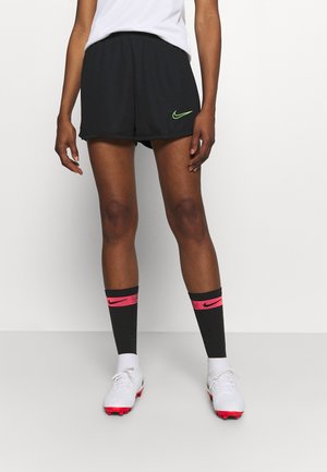 Persona con pantalones cortos negros de Nike con un logo verde, calcetas negras hasta la rodilla con rayas rosas y zapatillas blancas de fútbol, de pie contra una pared blanca.