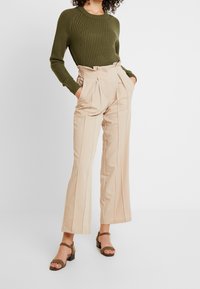 Beige Hochbundhosen mit Falten, schmal zulaufendem Bein und vertikalen Linien. Ein olivgrüner Rippenpullover und braune Absatzsandalen runden das Outfit ab.