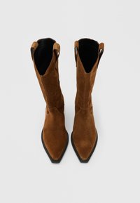 Ζεύγος καφέ σουέντ καουμπόικων μπότες με μυτερές μύτες και μαύρες σόλες, σε θέαση από πάνω σε απλό ανοιχτόχρωμο φόντο.