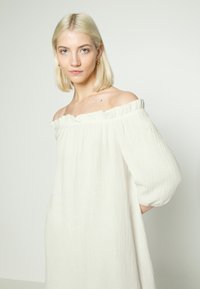 Vestido off-shoulder de cor creme com decote franzido e tecido texturizado. Apresenta mangas volumosas e uma silhueta descontraída.