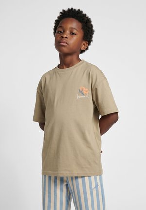 Dreng med krøllet hår iført en beige t-shirt med et lille blomstret logo og blå-hvidstribede bukser, står med hænderne bag ryggen.