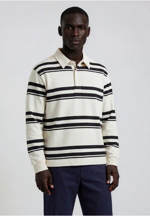 Homme portant un maillot de rugby blanc à manches longues avec des rayures horizontales noires et un pantalon bleu marine, debout devant un fond uni.