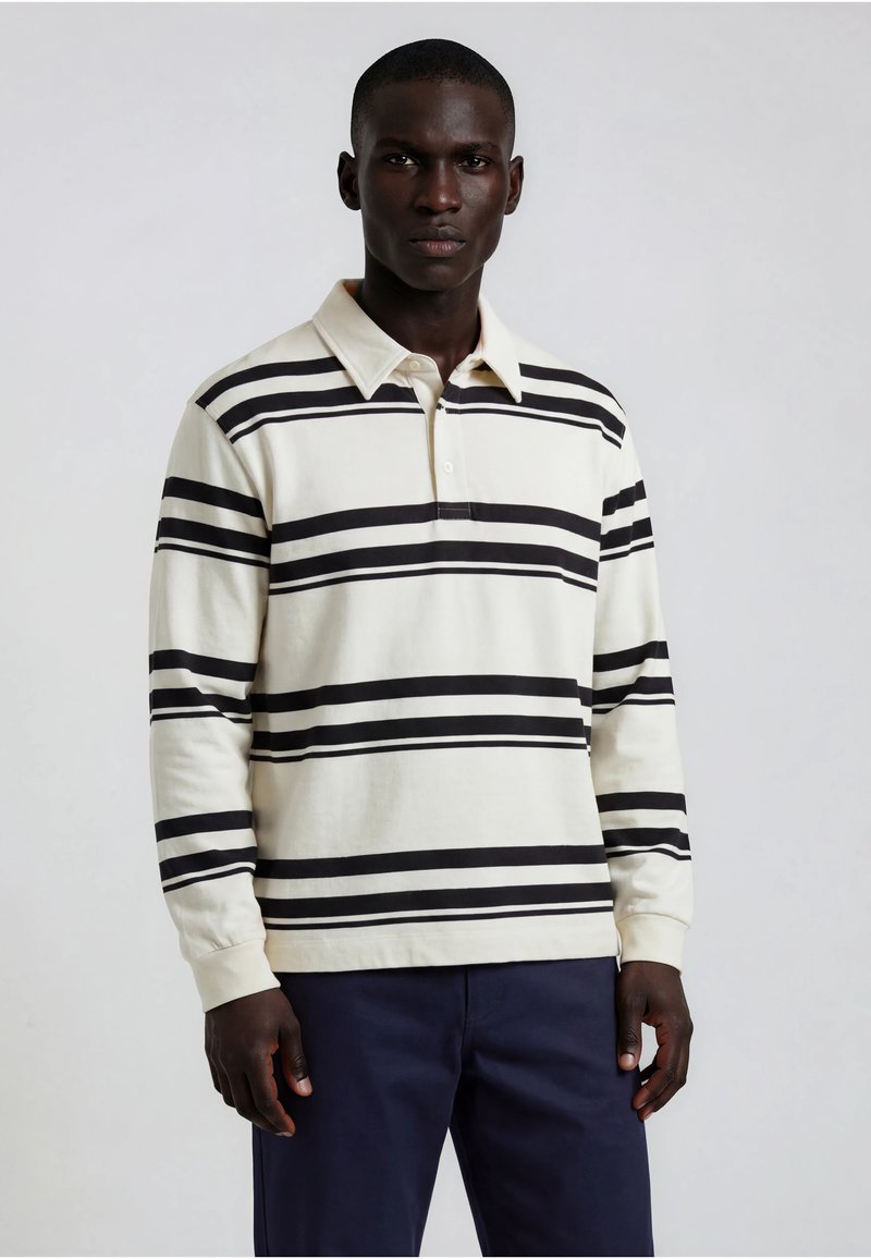 Homme portant un maillot de rugby blanc à manches longues avec des rayures horizontales noires et un pantalon bleu marine, debout devant un fond uni.