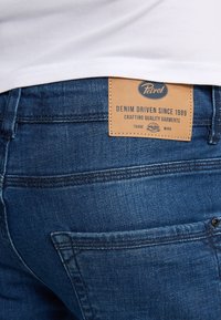 Jeans in denim blu scuro con una finitura testurizzata. Presentano un'etichetta in pelle con la scritta "Petrol" e cuciture a vista lungo le tasche e le cuciture.