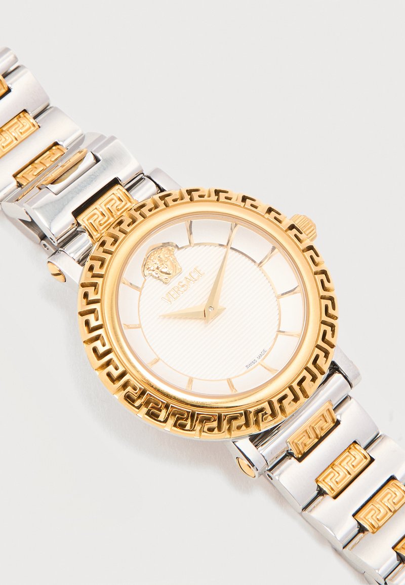 Montre-bracelet ronde en argent et or Versace avec logo Méduse, motif clé grec sur la lunette et le bracelet, affichant l'heure 10h02.