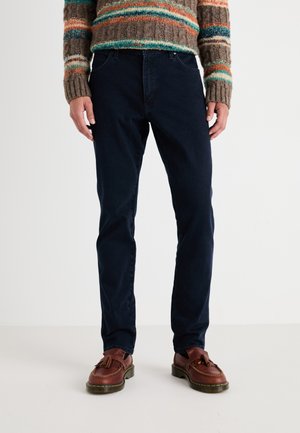 Donkerblauwe jeans in een slim fit ontwerp, met een klassieke vijf-pocket stijl en subtiele stiksels. Gecombineerd met bruin leren schoenen.