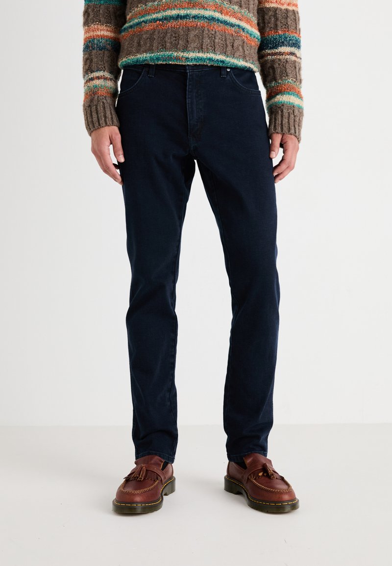 Vaqueros azul marino en un diseño ajustado, con un estilo clásico de cinco bolsillos y costuras sutiles. Combinados con zapatos de cuero marrón.