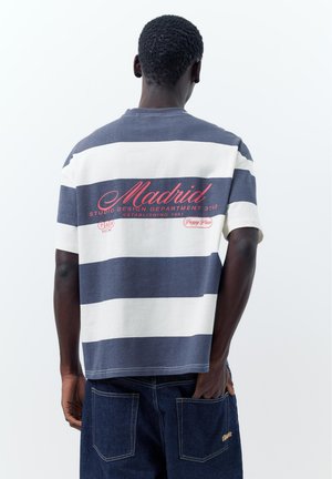 Háttal álló férfi, kék-fehér csíkos pólót visel "Madrid" felirattal, sötétkék farmernadrágban, kezét a hátsó zsebében tartja.