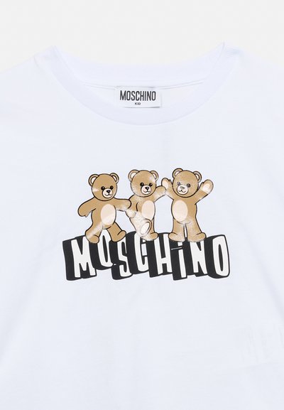 Három barna plüssmackó fehér hassal, fekete-fehér stilizált "MOSCHINO" felirat felett, fehér gyerekpólón.
