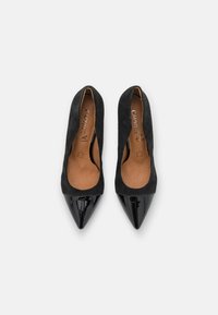 Escarpins en suede noir à talon haut avec une pointe en cuir verni brillant. L'intérieur présente une semelle en tan avec un logo. Design élégant et minimaliste.