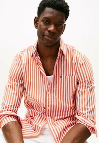Camicia a righe con strisce verticali arancioni e bianche, in cotone, con una tasca sul petto, maniche arrotolate e una consistenza morbida.