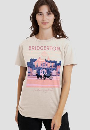 Young woman wearing beige Bridgerton t-shirt with pink and blue illustration of historic mansion and horse-drawn carriage in London, England.