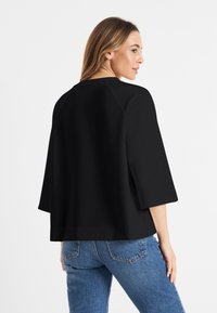 Vrouw in een losse zwarte top met mouwen tot de elleboog en blauwe jeans, iets naar de zijkant gekeerd tegen een effen witte achtergrond.