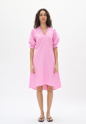 Pink linen dress with a V-neck, puff sleeves, and ruffled detailing. Flowy design with a slightly longer back hem. Worn with black sandals.