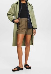 Vihreä trench-kurta, musta yläosa, farkkupaido, khaki-shortsit ja mustat sandaalit. Ominaisuuksiin kuuluu rento muotoilu ja erilaiset pinnat.