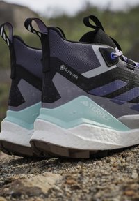 Paire de chaussures de randonnée Gore-Tex Terrex noires, violettes et bleu clair, posées sur un sol rocheux avec des gouttes d'eau à la surface.